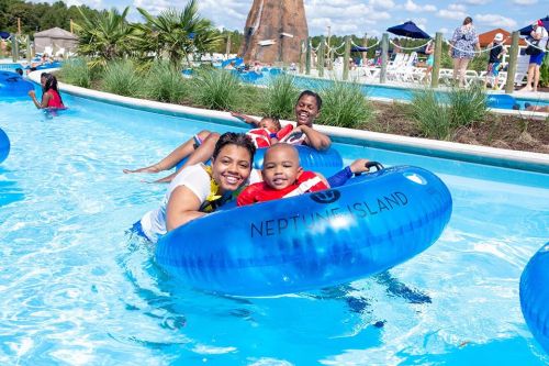 A woman and a child smile while floating in a blue inner tube in a water park pool, surrounded by greenery and other guests.