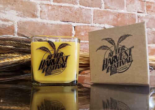 A yellow candle in a glass jar next to a kraft box, both featuring "After Harvest Festival" branding, with a brick background.