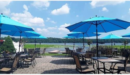 Outdoor dining area with blue umbrellas overlooking a lake, surrounded by greenery and a clear blue sky.