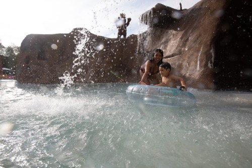 A woman and a boy enjoy a water slide, splashing in a pool with a rocky backdrop and a waterfall.