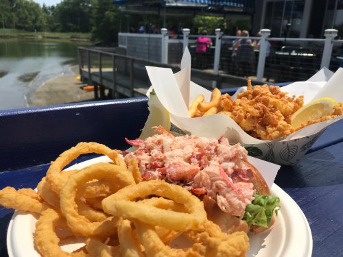A lobster roll with lettuce, onion rings, and a side of fried seafood, set by a waterfront dining area.