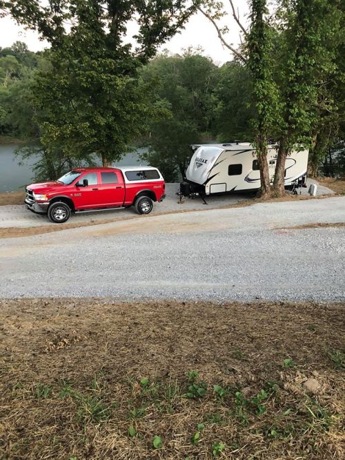 A red pickup truck parked next to a white travel trailer by a river, surrounded by trees and gravel.