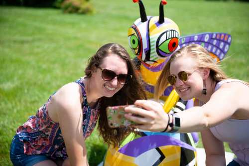 Two young women take a selfie with a colorful insect sculpture in a grassy outdoor setting.