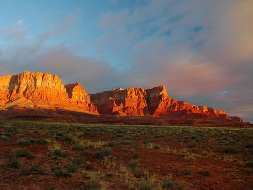 Sunset over rugged red rock formations, casting warm hues on the landscape with scattered grass in the foreground.