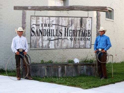 Two cowboys in hats and western attire stand beside the Sandhills Heritage Museum sign, holding ropes.