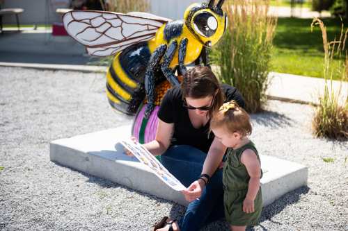 A woman and a toddler examine a colorful bee sculpture in a park, surrounded by greenery and gravel.