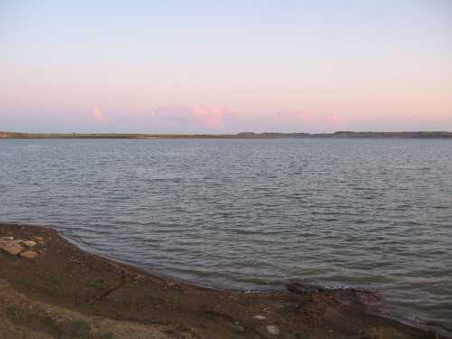 A serene lake at dusk, with calm waters reflecting a pastel sky and distant hills on the horizon.