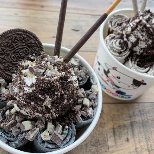 A bowl of rolled ice cream topped with whipped cream, crushed Oreos, chocolate drizzle, and Pocky sticks.