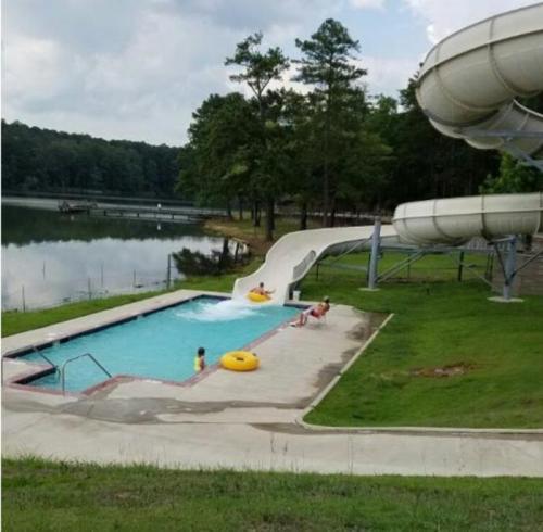 A water slide next to a pool by a lake, with children playing and lounging on inflatable rings. Trees in the background.