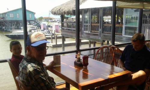 A man and a boy sit at a table in a waterfront restaurant, with a view of boats and a thatched-roof building outside.