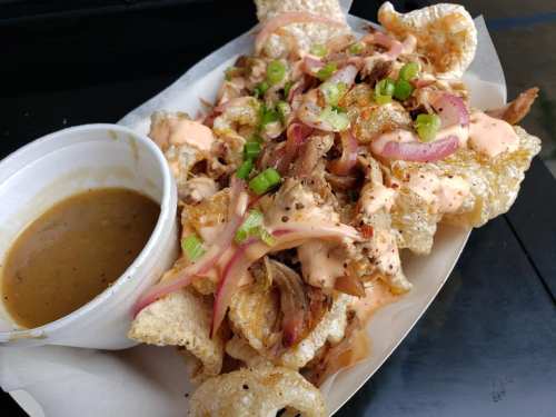 A plate of crispy pork rinds topped with pulled meat, pink onions, green onions, and a side of sauce.