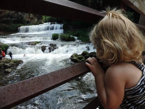 A child with curly hair gazes at a waterfall from behind a railing, with people enjoying the water below.
