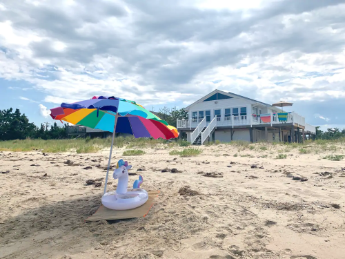 A colorful beach umbrella shades a unicorn float on the sand, with a beach house in the background under a cloudy sky.