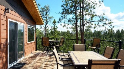 A wooden deck with outdoor furniture overlooks a forested area under a blue sky with scattered clouds.