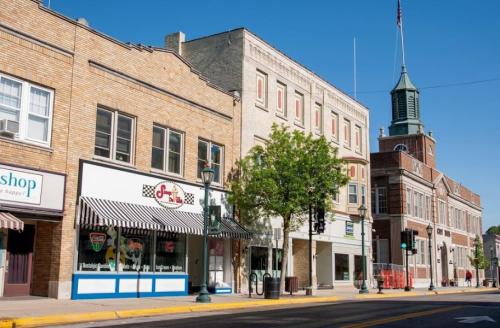 A sunny street view featuring brick buildings, a shop with striped awning, and a clock tower in the background.