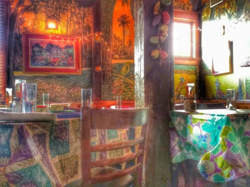 Cozy restaurant interior with colorful tablecloths, wooden chairs, and vibrant wall art featuring nature scenes.