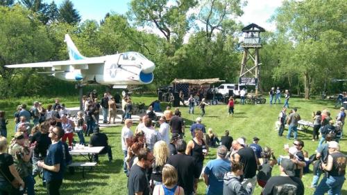 A lively outdoor event with a crowd gathered around a stage, featuring a suspended airplane and a lookout tower in the background.