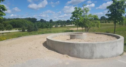 A circular concrete structure filled with sand, surrounded by greenery and blue skies with scattered clouds.