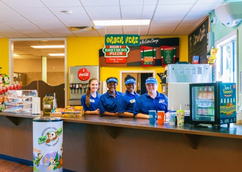 Four employees in blue uniforms smile behind a counter at a convenience store, with snacks and drinks displayed.