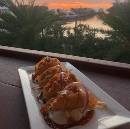 A plate of tacos topped with sauce and onions, set against a sunset view over a marina.