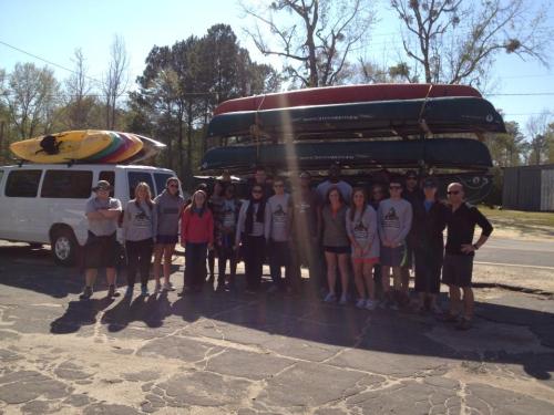 A group of people stands in front of a van with stacked kayaks and canoes, ready for an outdoor adventure.