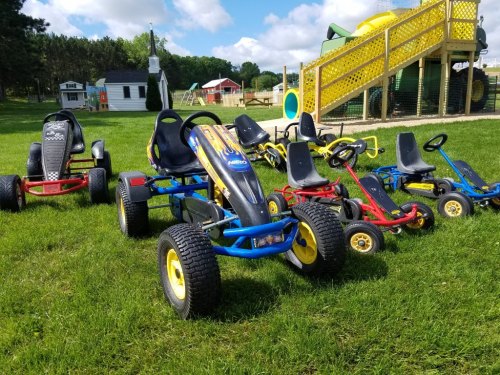 A row of colorful go-karts parked on green grass near a playground with slides and play structures in the background.