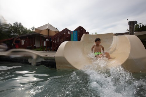 A child slides down a water slide into a pool, creating a splash, with a sunny outdoor setting in the background.