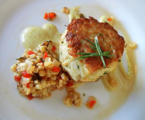 A plated dish featuring a crab cake, creamy sauce, and a side of corn salad with diced peppers.