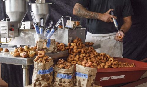 A chef prepares mini donuts, surrounded by bags of various flavors and a tray filled with freshly made donuts.