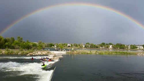 A vibrant rainbow arcs over a river with kayakers navigating the rapids, surrounded by greenery and a cloudy sky.