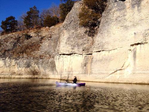 A person kayaking on calm water near a rocky cliff under a clear blue sky.