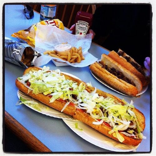 A table with a large sub sandwich topped with lettuce, a plate of fries, and another sandwich in the background.