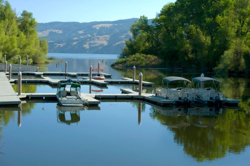 A serene lake scene with boats docked at wooden piers, surrounded by lush greenery and calm water reflecting the landscape.