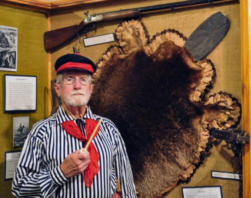 An elderly man in period clothing holds a pipe, standing in front of a fur hide and historical weapons display.