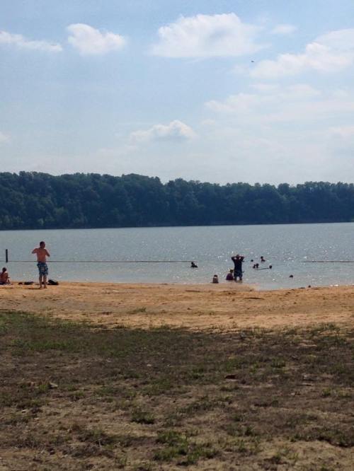 A sandy beach with people swimming and playing in the water under a sunny sky with scattered clouds.