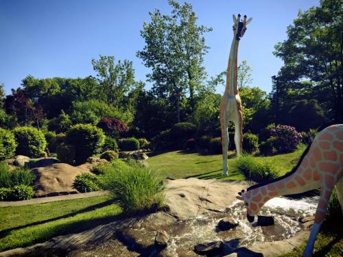 Two large giraffe sculptures in a lush garden with greenery and a small stream under a clear blue sky.