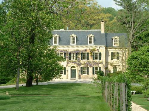 A large, elegant house with a slate roof, surrounded by lush greenery and blooming flowers in a serene landscape.
