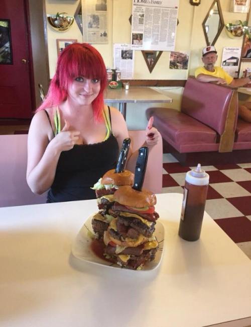 A woman with red hair gives a thumbs up next to a towering burger stack with two knives inserted, in a diner setting.