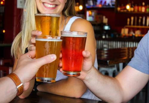 Three people clink glasses of beer in a lively bar setting, smiling and enjoying their drinks.