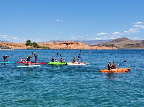 A group of people kayaking on a sunny day, with mountains and blue sky in the background.