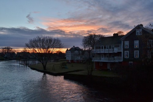 A serene river scene at sunset, featuring houses along the bank and a colorful sky with clouds.