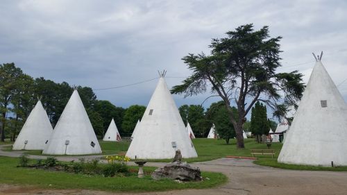 A cluster of white teepee-shaped structures in a grassy area, surrounded by trees and a cloudy sky.