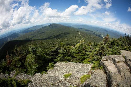 A panoramic view from a rocky summit, showcasing lush green mountains and a blue sky with scattered clouds.
