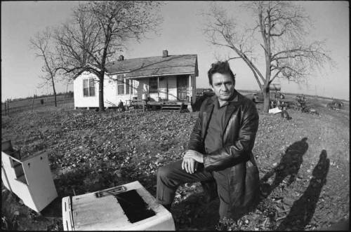 A man in a leather jacket kneels beside an old appliance in front of a rural house, surrounded by barren trees.