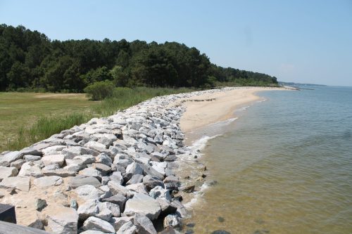 A rocky shoreline curves along a calm bay, bordered by green trees and sandy beach under a clear blue sky.