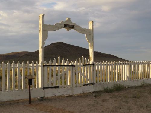 A weathered white gate labeled "Cemetery" opens to a barren landscape with a distant hill under a cloudy sky.