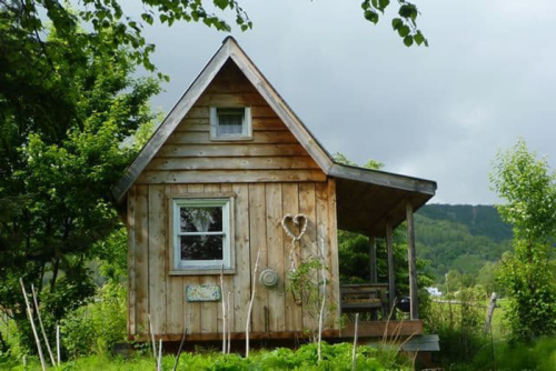 A small wooden cabin with a sloped roof, surrounded by greenery and trees under a cloudy sky.