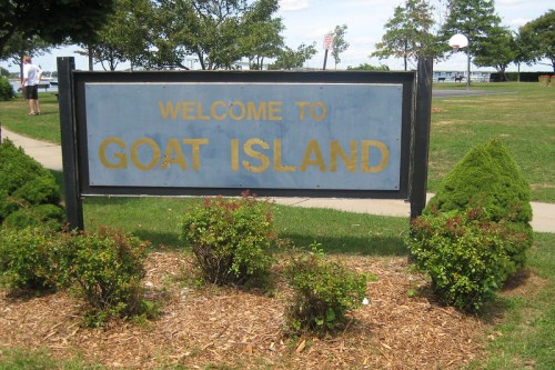 Sign reading "Welcome to Goat Island" surrounded by greenery and a pathway, with trees and a basketball court in the background.