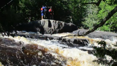Two people stand on a rock by a rushing river, surrounded by lush green trees and cascading water.