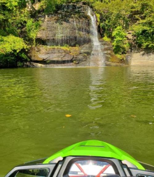 A green jet ski in the foreground with a serene lake and a waterfall surrounded by lush greenery in the background.